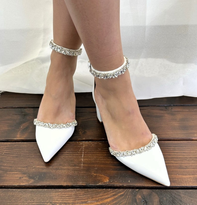 May include: White satin bridal shoes with a pointed toe and ankle strap adorned with crystal embellishments.