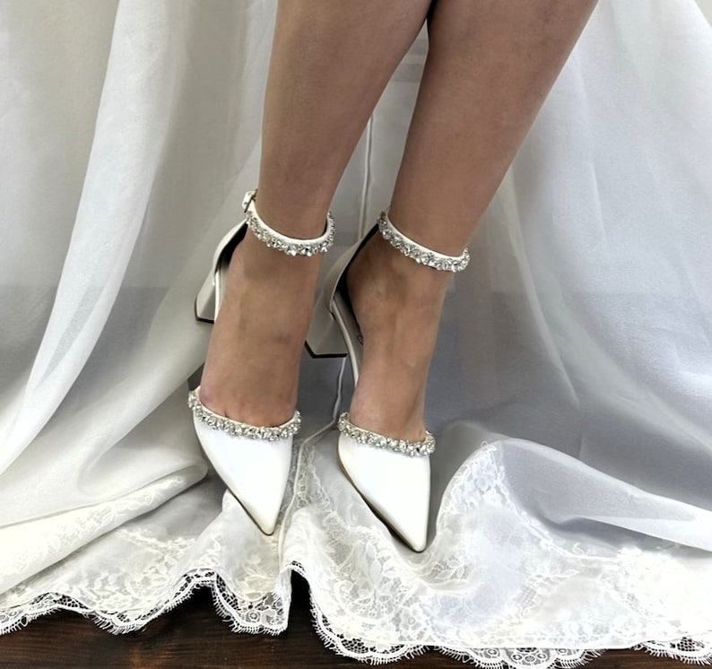 May include: White satin bridal heels with a pointed toe and ankle strap adorned with crystal embellishments.