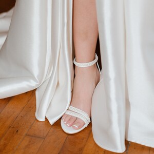 Puede incluir: Vestido de novia de sat&eacute;n blanco con una abertura en la falda, que muestra una sandalia blanca de tac&oacute;n con adornos de perlas.