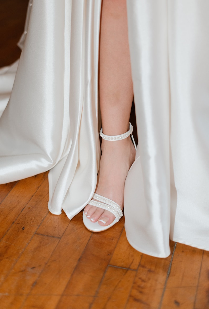 Puede incluir: Vestido de novia de sat&eacute;n blanco con una abertura en la falda, que muestra una sandalia blanca de tac&oacute;n con adornos de perlas.