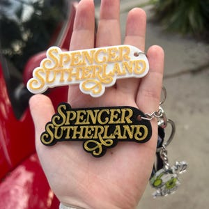 May include: Two keyrings with the name "Spencer Sutherland" in a decorative font. One is white with gold lettering, the other black with gold lettering. Both have key rings attached.