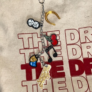May include: A silver chain with charms attached. The charms include a pair of dice, a horseshoe, a cherry, a musical note, a pair of theater masks, and a gold charm with the text "The Greatest Show".