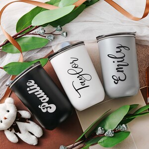 May include: Three personalized travel mugs with names 'Sophie', 'Emily', and 'Taylor' written in stylish script. The mugs are in black, gray, and white, respectively.  They appear to be stainless steel or similar material with a sleek, modern design. The mugs are displayed on a surface with greenery and ribbon, suggesting a gift or celebratory theme.