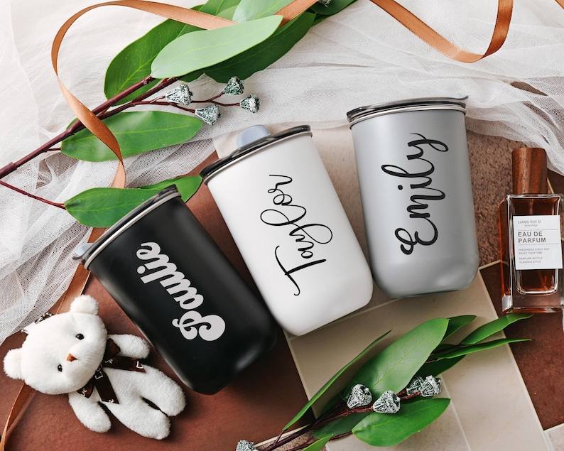 May include: Three personalized travel mugs with names 'Sophie', 'Emily', and 'Taylor' written in stylish script. The mugs are in black, gray, and white, respectively.  They appear to be stainless steel or similar material with a sleek, modern design. The mugs are displayed on a surface with greenery and ribbon, suggesting a gift or celebratory theme.