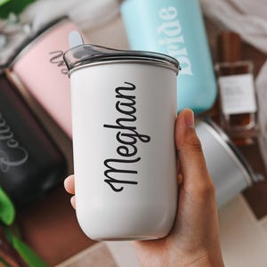 May include: Close-up view of several personalized stainless steel travel tumblers.  Each tumbler features a different name, including 'Colleen', 'Bride', and 'Meghan', engraved in a stylish script font. The tumblers are in various colors: black, pink, light blue, and white. The tumblers appear to be insulated and have lids.  The white tumbler is prominently displayed in the center, showcasing the name 'Meghan'.