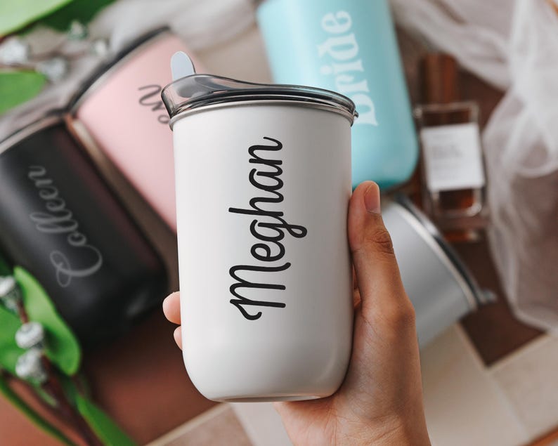 May include: Close-up view of several personalized stainless steel travel tumblers.  Each tumbler features a different name, including 'Colleen', 'Bride', and 'Meghan', engraved in a stylish script font. The tumblers are in various colors: black, pink, light blue, and white. The tumblers appear to be insulated and have lids.  The white tumbler is prominently displayed in the center, showcasing the name 'Meghan'.