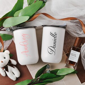 May include: Two white personalized insulated stainless steel tumblers with black lids. One tumbler says 'Audrey' in a pink script font, and the other says 'Danielle' in a black script font.  A small white teddy bear and a brown bottle of perfume are also in the image. The tumblers are displayed on a brown surface with green leaves and a brown ribbon.