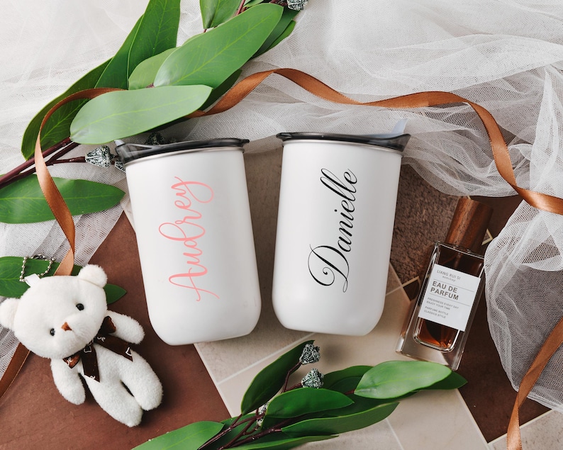 May include: Two white personalized insulated stainless steel tumblers with black lids. One tumbler says 'Audrey' in a pink script font, and the other says 'Danielle' in a black script font.  A small white teddy bear and a brown bottle of perfume are also in the image. The tumblers are displayed on a brown surface with green leaves and a brown ribbon.