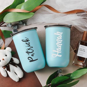May include: Two light blue personalized stainless steel travel mugs with lids and handles. One mug says 'Patrick' and the other says 'Hannah'.  A small stuffed white teddy bear and a brown bottle of perfume are also in the image.