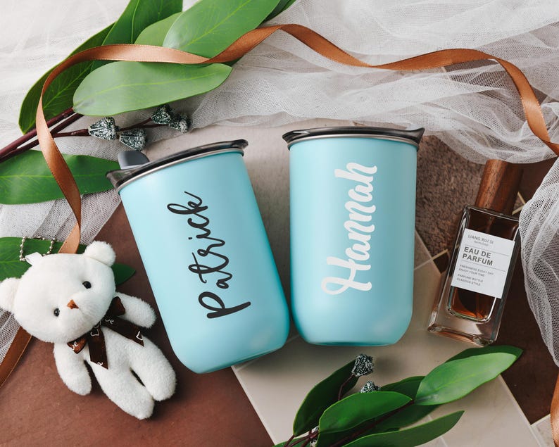 May include: Two light blue personalized stainless steel travel mugs with lids and handles. One mug says 'Patrick' and the other says 'Hannah'.  A small stuffed white teddy bear and a brown bottle of perfume are also in the image.