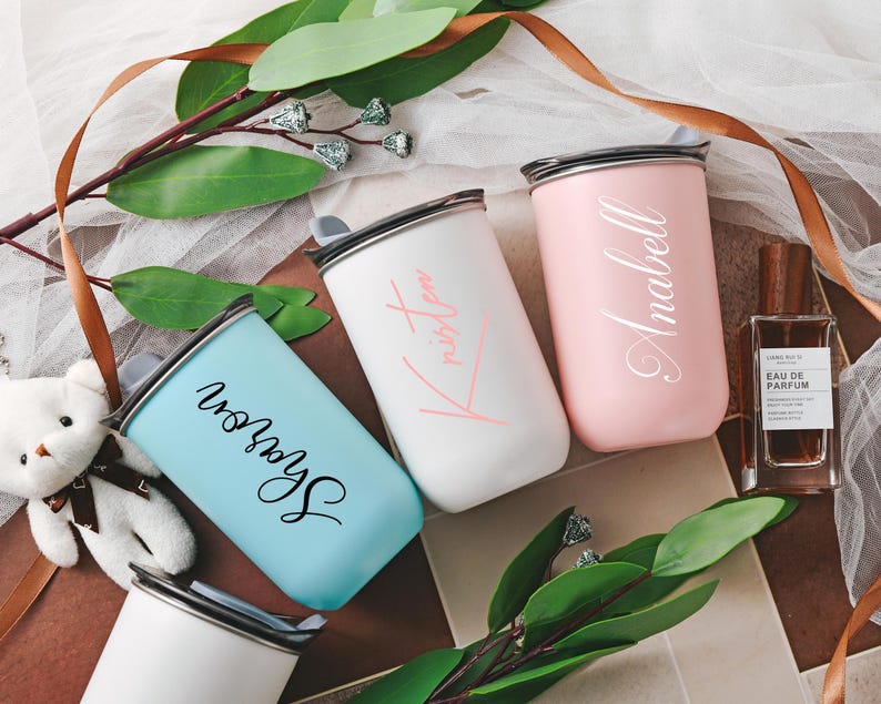 May include: Three personalized pastel-colored stainless steel tumblers with names 'Anabell', 'Kristen', and 'Sharon' inscribed in elegant script.  The tumblers are light pink, white, and light blue. They feature a modern, minimalist design and are ideal for gifting or personal use. The tumblers are displayed on a surface with greenery and ribbon.
