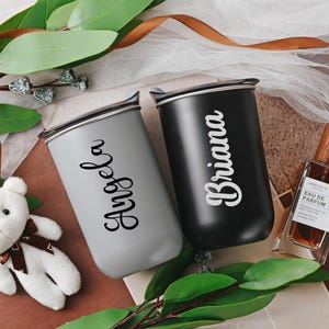 May include: Two personalized tumblers, one gray and one black, feature names 'Angela' and 'Briana' in a stylish script font.  The tumblers have a sleek, modern design and appear to be made of durable material.  A brown leather-like ribbon and a small stuffed white teddy bear are in the background. A bottle of perfume is also visible.