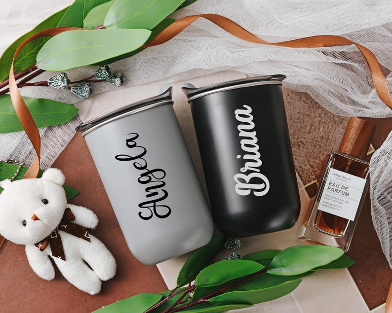 May include: Two personalized tumblers, one gray and one black, feature names 'Angela' and 'Briana' in a stylish script font.  The tumblers have a sleek, modern design and appear to be made of durable material.  A brown leather-like ribbon and a small stuffed white teddy bear are in the background. A bottle of perfume is also visible.