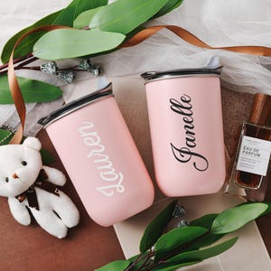 May include: Two pink personalized tumblers with the names Lauren and Janelle.  The tumblers have a metallic lid and are displayed with a small stuffed white bear, brown ribbon, and a bottle of perfume. The overall theme is a gift set or bridal shower.