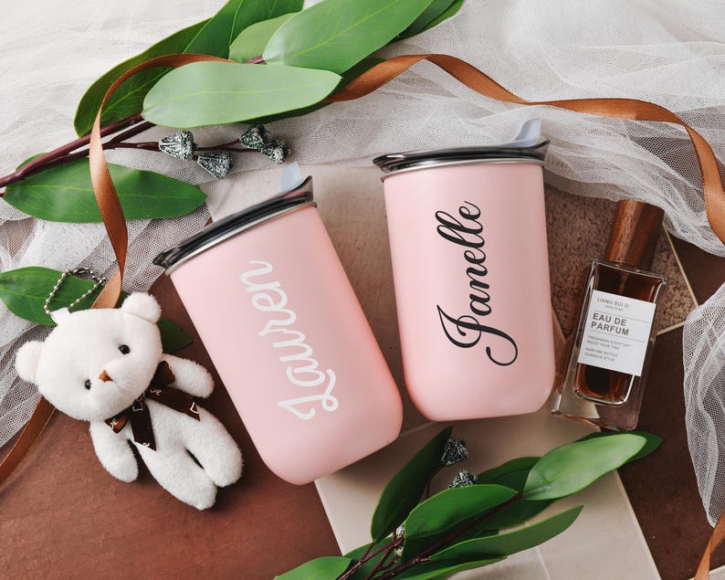 May include: Two pink personalized tumblers with the names Lauren and Janelle.  The tumblers have a metallic lid and are displayed with a small stuffed white bear, brown ribbon, and a bottle of perfume. The overall theme is a gift set or bridal shower.
