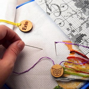 May include: Cross-stitch project in progress, with white fabric stretched in a blue hoop. A needle is threaded with purple thread. Wooden tags with the name "Erin" and an owl design are attached. A pattern chart is visible.
