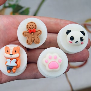 Può includere: Una collezione di quattro elementi decorativi bianchi e rotondi. Uno presenta un omino di pan di zenzero con un papillon rosso, un altro un panda con guance rosee, una volpe con una camicia blu e un'impronta di zampa rosa.