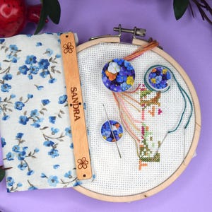 May include: A cross-stitch embroidery kit with a wooden hoop, needle, and floral fabric. The kit includes a wooden tool with the name "Sandra" and a floral pattern. The fabric has a blue floral design.