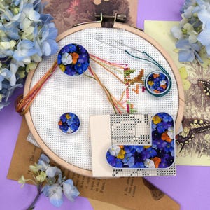 Peut inclure: Un cercle à broder avec des fils colorés, entouré d'éléments floraux et décoratifs. Le design comprend un motif de point de croix partiellement terminé et plusieurs ornements sur le thème floral, dont une épingle circulaire et une pièce décorative en forme de L.