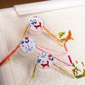 May include: Three white plastic bobbins with colorful floral and fox designs are used to hold embroidery floss on a white Aida cloth. The floss is threaded through the cloth and secured to the bobbins.