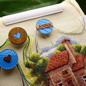 May include: Cross-stitch project in progress, featuring a detailed house scene. Wooden thread holders with heart cutouts and the name "ANNA" are visible. Colorful threads and a white frame complete the craft setup.