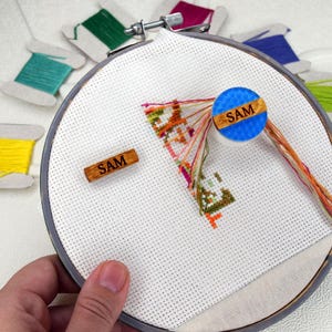 Peut inclure: Un projet de point de croix dans un cercle à broder, avec des fils colorés et un motif partiellement terminé. Le mot "SAM" est visible sur deux accents en bois. Des bobines de fil de différentes couleurs entourent le cercle, suggérant un thème artisanal.