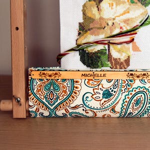 May include: A wooden embroidery frame holding a cross-stitch project with a floral design. The fabric is secured with colourful threads. The frame has a decorative fabric with a paisley pattern in teal, brown, and cream. The name "MICHELLE" is visible.