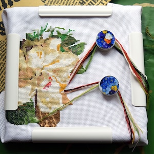 May include: White embroidery hoop with a cross-stitch design of a white flower with green leaves. The hoop is held in place by two white clips. Two round, colorful thread organizers are attached to the hoop with colorful threads.