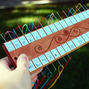 May include: A wooden needle minder with multiple needles threaded with colorful embroidery floss. The minder has a series of numbered squares with small diagrams and decorative swirls. The numbers include 221, 035, 036, 037, 222, 224, 228, 271, 233, 278, 252, 250, 260, 261, 223, and 288.