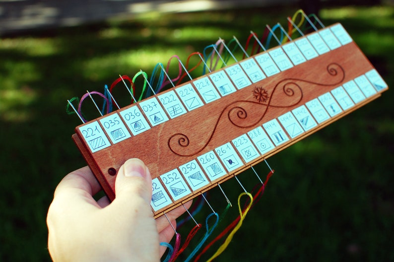 May include: A wooden needle minder with multiple needles threaded with colorful embroidery floss. The minder has a series of numbered squares with small diagrams and decorative swirls. The numbers include 221, 035, 036, 037, 222, 224, 228, 271, 233, 278, 252, 250, 260, 261, 223, and 288.