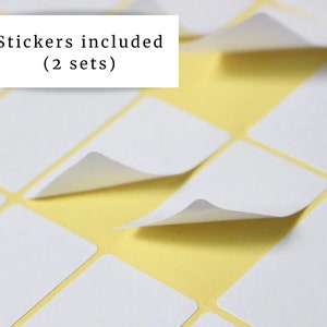 May include: Close-up of white rectangular stickers with a peel-off corner, arranged on a yellow background. The text "Stickers included (2 sets)" is in a white rectangle in the upper left corner. The stickers are arranged in a grid pattern.