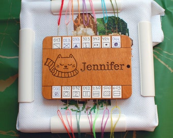 May include: A wooden cross-stitch thread organizer with the name "Jennifer" engraved, featuring a cat design. The organizer holds various colored threads and is set on a white fabric, with a green background.