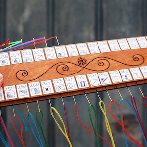 May include: A wooden thread organizer with multiple colored threads attached. The organizer has a series of numbered slots with small drawings. The wood is decorated with a swirl design and a snowflake. The organizer is held by a hand.