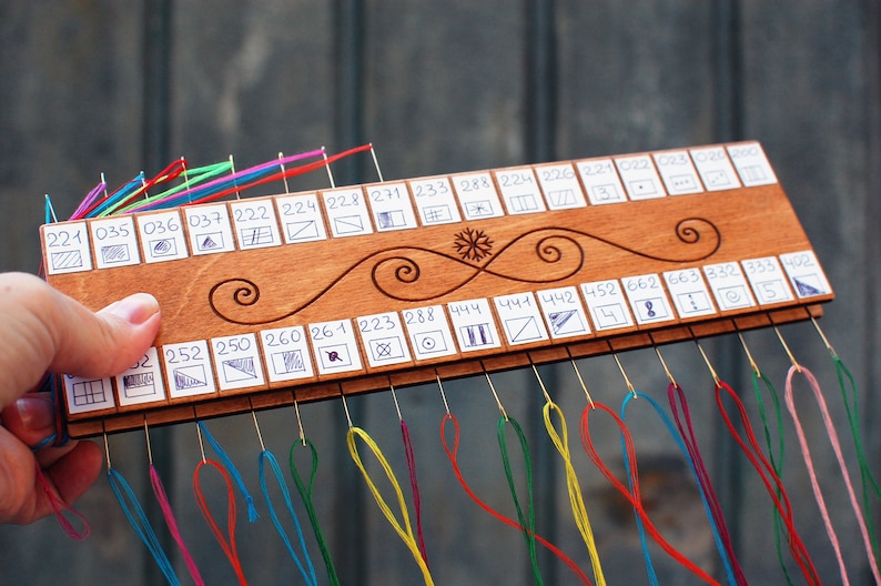 May include: A wooden thread organizer with multiple colored threads attached. The organizer has a series of numbered slots with small drawings. The wood is decorated with a swirl design and a snowflake. The organizer is held by a hand.