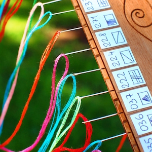 May include: Close-up of a wooden embroidery frame with colorful threads and needles. The frame has small white labels with numbers and simple drawn patterns. The threads are in shades of red, pink, blue, green, and yellow.