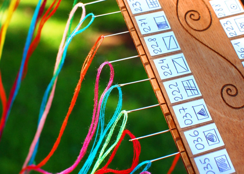 May include: Close-up of a wooden embroidery frame with colorful threads and needles. The frame has small white labels with numbers and simple drawn patterns. The threads are in shades of red, pink, blue, green, and yellow.