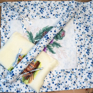 May include: A white apron with blue floral print and matching ties, featuring a cross-stitched design of a green and purple thistle. Additional cross-stitch pieces include a house and a small pillow, all on a floral background.