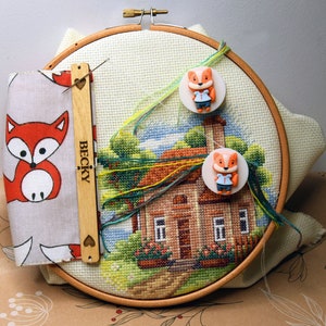 May include: Cross-stitch embroidery hoop with a completed house scene. Includes a fabric case with a fox design, wooden needle minder with "BECKY" text, and two fox-shaped button magnets. The embroidery features a house, trees, and a blue sky.