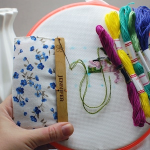 Può includere: Un progetto di punto croce in corso, con tessuto bianco teso in un telaio rotondo. Sono visibili fili colorati per ricamo nei toni del rosa, giallo, verde e blu. È presente anche un porta aghi in legno con il nome "JENNIFER".