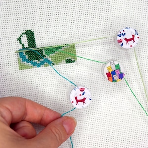 Op de afbeelding: Een kruissteekproject in uitvoering, met een gedeeltelijk gestikt groen en blauw ontwerp op witte stof. Drie ronde naaldenhouders met illustraties van vossen en katten zijn met draad aan de stof bevestigd en houden de naald op zijn plaats.