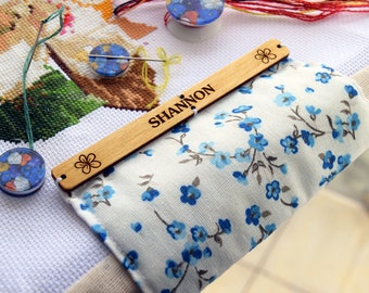 May include: A wooden needle minder with the name "SHANNON" engraved, alongside a floral design. The minder rests on a floral fabric, with a cross-stitch project in the background. Other craft supplies are also visible.