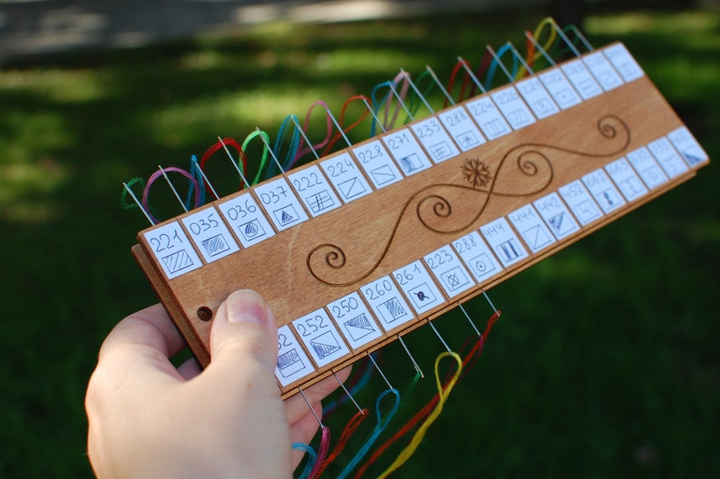 May include: A wooden embroidery floss organizer with multiple needles and colored threads. The organizer has a series of numbered slots with small diagrams. The wood is light brown with decorative swirls. The threads are in various colors.