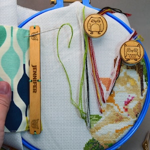Pode incluir: Um projeto de ponto cruz num bastidor azul, com um desenho floral parcialmente concluído. Um porta-agulhas de madeira com o nome "JENNIFER" e um desenho de coruja está fixado. Porta-agulhas de madeira em forma de coruja e fios coloridos também são visíveis.