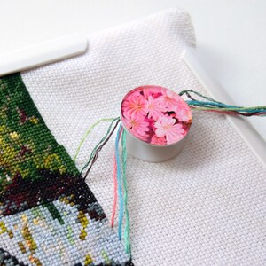 May include: A white plastic embroidery thread organizer with a pink floral design. The organizer has multiple slots for holding different colors of embroidery floss.