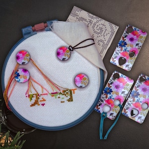 May include: A cross-stitch embroidery hoop with a partially completed design, colorful threads, and floral-patterned magnets. Three phone cases with a matching floral design are also present, along with a piece of fabric and a pattern.