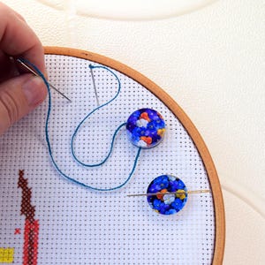 Puede incluir: Primer plano de un proyecto de punto de cruz en un aro de bordado de madera. El proyecto presenta un diseño colorido con flores azules y naranjas. Dos cuentas florales azules y naranjas están unidas a la tela.