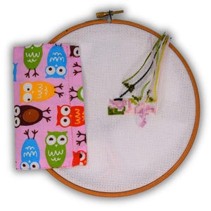 May include: A pink fabric with a pattern of colorful owls is wrapped around a wooden embroidery hoop with white Aida cloth. The hoop is empty except for some loose embroidery floss.