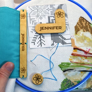 May include: Cross-stitch embroidery hoop with a partially completed floral design. Includes wooden tools with the name "JENNIFER" engraved, a teal fabric needle holder, and colorful embroidery floss. A needle is threaded with blue floss.