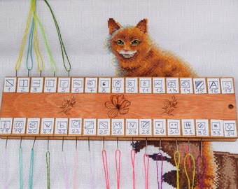 Peut inclure: Un organiseur d'aiguilles en bois avec des emplacements numérotés et des symboles, tenant des aiguilles avec des fils colorés. Une broderie au point de croix d'un chat orange est en arrière-plan. L'organisateur d'aiguilles a un motif floral.