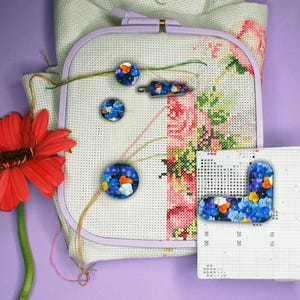 Peut inclure: Un projet de point de croix dans un cercle de broderie lilas, avec broderie florale. Plusieurs aimants décoratifs avec des motifs floraux bleus et oranges sont éparpillés. Un gerbera rouge est à gauche.